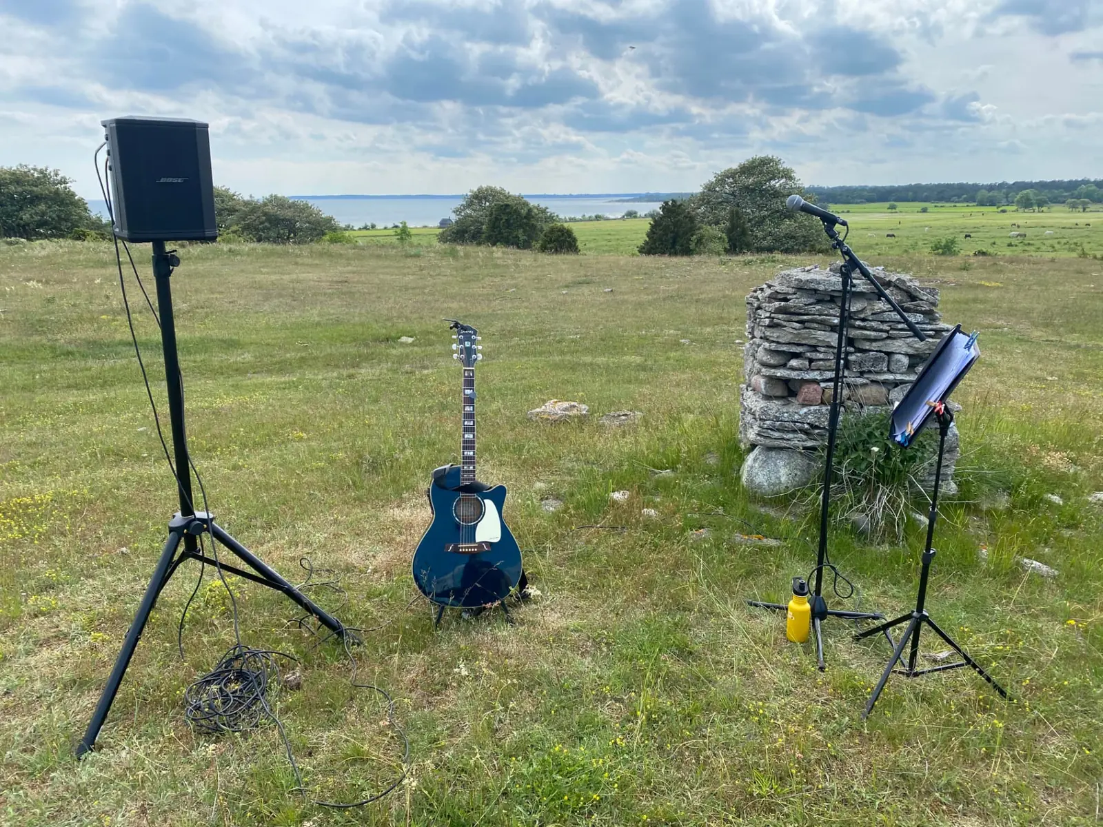 Musikutrustning med högtalare, akustisk gitarr och mikrofon uppställd på en gotländsk äng med utsikt över havet